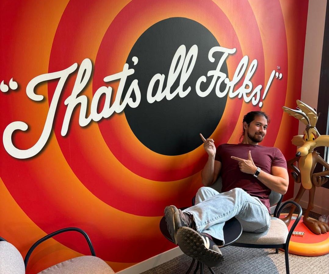 Nicholas Biggs, feet up on a café table, pointing at a giant “That's all Folks!” wall, with Wile E. Coyote looking on